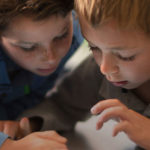 Zwei Jungen liegen auf dem Boden, betrachten etwas außerhalb des Bildes und interagieren damit. Sie wirken konzentriert und interessiert. Einer trägt ein blau gestreiftes Hemd, der andere ein graues Poloshirt.