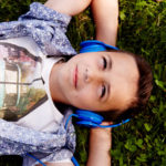 Ein Junge mit kurzen braunen Haaren liegt im grünen Gras. Er trägt ein gemustertes Hemd, ein T-Shirt mit Autoaufdruck und blaue Kopfhörer. Er blickt entspannt nach oben, die Arme hinter dem Kopf verschränkt.