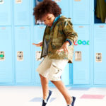 Ein kleines Kind in Tarnjacke, Shorts und Turnschuhen tanzt fröhlich vor einer Reihe hellblauer Schließfächer und schreibt mit jeder unbekümmerten Bewegung Geschichte.