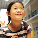 Ein junges Mädchen mit Zöpfen und gestreiftem Hemd lächelt, während es in einem hellen, farbenfrohen Schulgebäude mit Kunstwerken an den Wänden, einer hohen Decke und Räumen für den Sportunterricht sitzt.