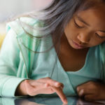 Ein junges Mädchen mit langem dunklem Haar und einem mintgrünen Pullover sitzt konzentriert an einem Schreibtisch, während sie mit ihrem Finger ein Tablet bedient. Dabei erkundet sie möglicherweise interaktive Sportaktivitäten oder informiert sich online über Sportunterricht.