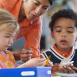 Eine Frau hilft zwei kleinen Kindern, einem Mädchen und einem Jungen, bei einer Bastelaktivität an einem Tisch. Die Kinder sind konzentriert beim Papierschneiden, während die Frau ihnen Anleitung gibt.