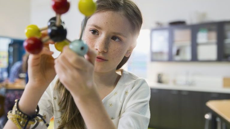 Ein junges Mädchen untersucht in einem Klassenzimmer ein buntes Molekülmodell und konzentriert sich ganz intensiv, während sie die verbundenen Kugeln hält und betrachtet.