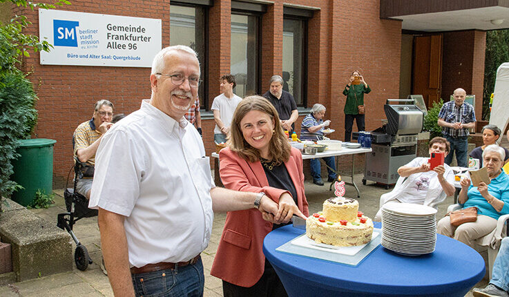 Ein lächelnder Mann und eine lächelnde Frau schneiden bei einer Feier im Freien in Hof anlässlich der 5-Jahres-Feier gemeinsam eine Torte an. Die Leute sitzen und schauen zu, einige machen Fotos. Im Hintergrund sind ein Stapel Teller und ein Schild mit der Aufschrift Gemeinde Frankfurter Allee 96 zu sehen.