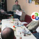 Ein Mann steht und präsentiert einer kleinen Gruppe, die um einen Tisch mit Getränken und Tellern in einer entspannten EinLaden-Einstellung sitzt. Einige Gesichter sind durch bunte Logos verdeckt, die dieser Szene voller Kunstwerke und einer Motivationsschub-Atmosphäre eine spielerische Note verleihen.