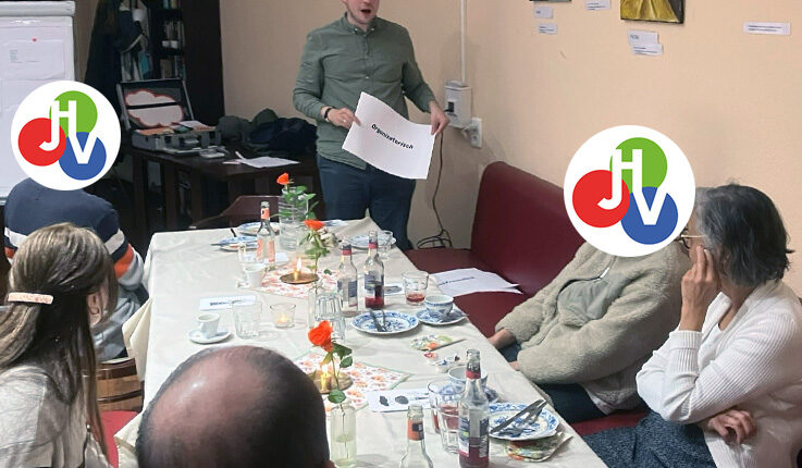 Ein Mann steht und präsentiert einer kleinen Gruppe, die um einen Tisch mit Getränken und Tellern in einer entspannten EinLaden-Einstellung sitzt. Einige Gesichter sind durch bunte Logos verdeckt, die dieser Szene voller Kunstwerke und einer Motivationsschub-Atmosphäre eine spielerische Note verleihen.