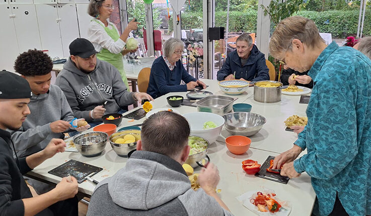Eine bunt gemischte Gruppe von Menschen, jung und alt, versammelt sich um einen Tisch und bereitet in einem hellen Raum mit großen Fenstern und viel Grün draußen gemeinsam Gemüse zu - eine Atmosphäre voller Wärme, ganz wie ein automatisch gespeicherter Entwurf von Zusammengehörigkeit.