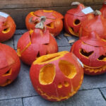 Mehrere geschnitzte Kürbisse mit gruseligen Gesichtern sind auf Steinplatten im Außenbereich der BUSSTOP Familienzentrum Halloween Party ausgestellt, jeder mit einem einzigartigen Gesichtsausdruck. Bei den orangefarbenen Kürbissen sind die Spitzen schräg aufgesetzt.