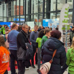 Eine geschäftige Indoor-Veranstaltung auf der ABI-Zukunft-Berlin in einem modernen Glasgebäude, mit Menschen, die sich unterhalten und Messestände besuchen. Einige Teilnehmer tragen grüne und orangefarbene Hemden mit Logos, und überall sind Informationsdisplays und Banner zu sehen.