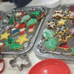 Drei Tabletts mit dekorierten Weihnachtsplätzchen - Sterne, Bäume und Tiere - schmücken den Tisch mit buntem Zuckerguss und Streuseln. Ein roter Teller und sternförmige Ausstechformen schaffen eine festliche Szene, perfekt für ein lebendiges Adventskalendertreffen.