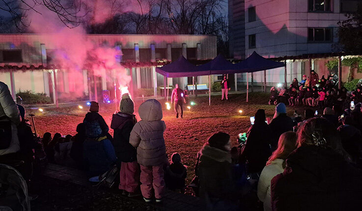 Auf dem Wintermarkt 2025 versammelt sich eine Menschenmenge, um eine nächtliche Feuervorstellung im Freien zu verfolgen. Die Darsteller blenden in der Mitte mit Rauch und Flammen, während die Zuschauer auf der Wiese sitzen und stehen. Gebäude und Zelte sind im Hintergrund beleuchtet.