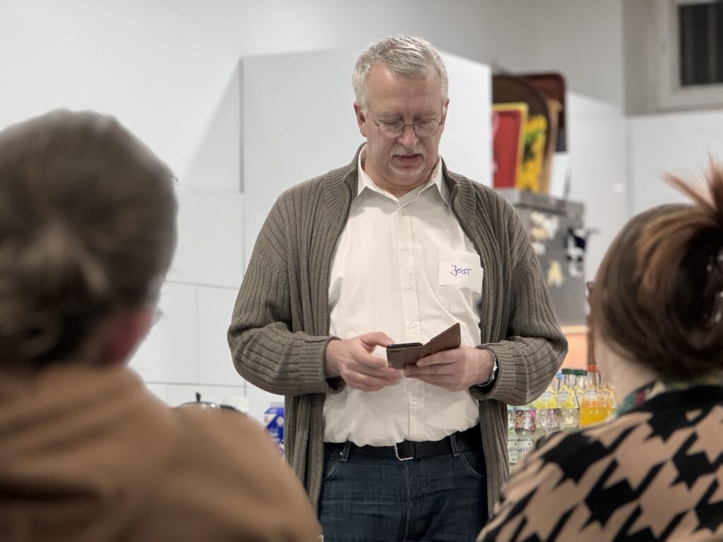 Ein Mann mit grauem Haar, Brille und brauner Strickjacke steht in einem Haus und liest in einem braunen Notizbuch. Zwei Teammitglieder sitzen im Vordergrund und hören zu, während sie gemeinsam feiern. Auf dem Tresen hinter ihm sind Flaschen und Getränke zu sehen.