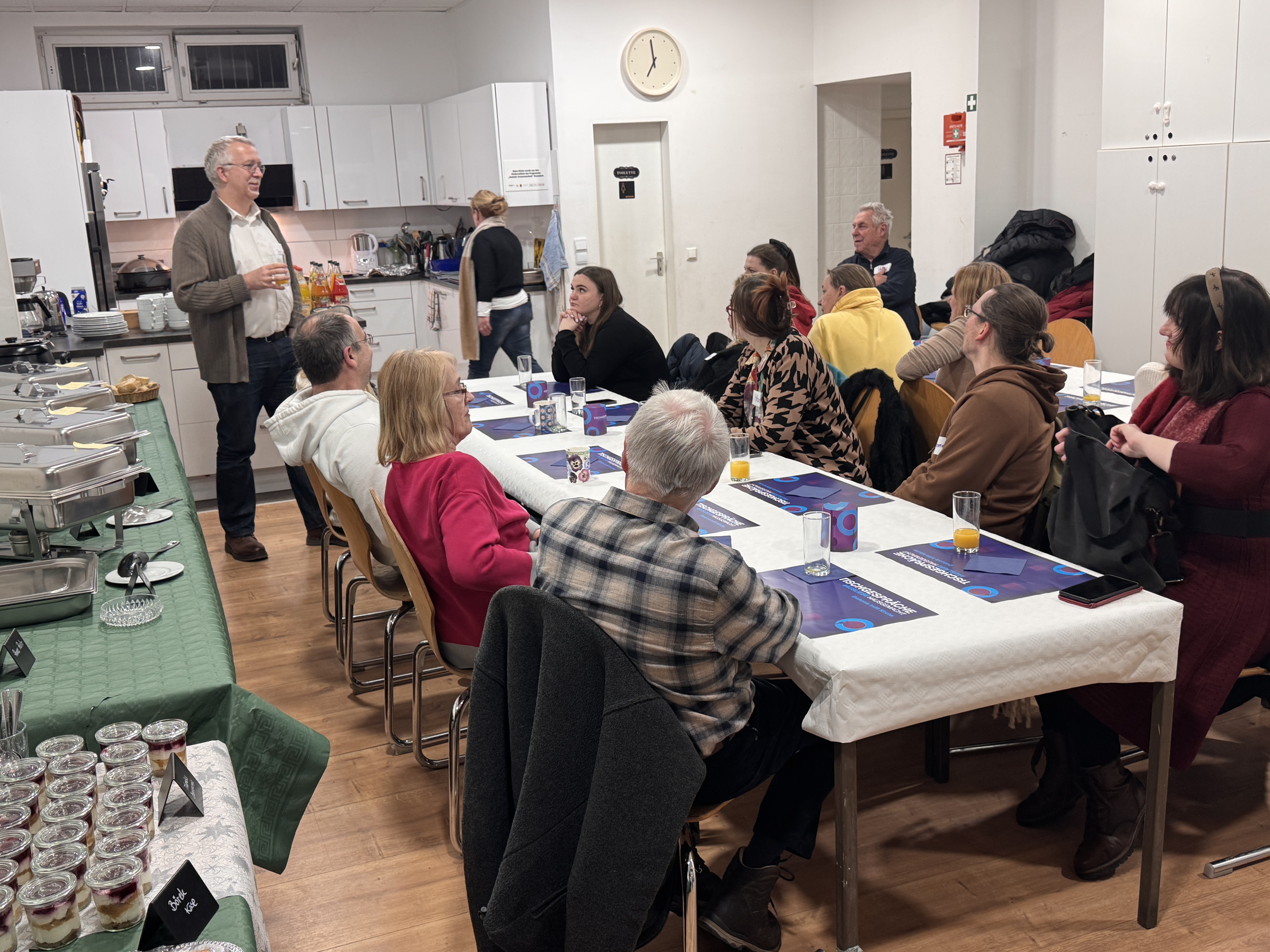 Eine Gruppe von Menschen sitzt um Tische in einer Küche oder einem Essbereich und genießt Speisen und Getränke, während ein Mann eine Rede hält. Die Atmosphäre ist lebhaft, wie geschaffen für eine Teamfeier mit Tischsets auf den Tischen und einem Buffet im Vordergrund.