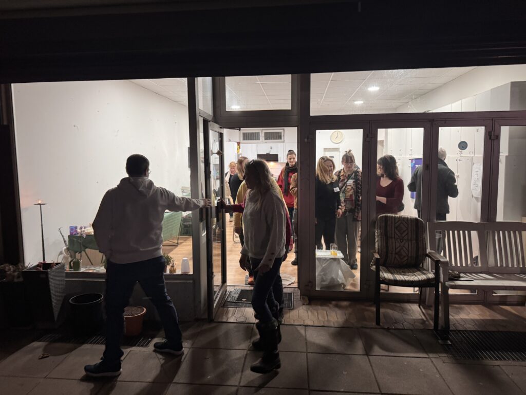 Ein Team versammelt sich in einem hell erleuchteten Raum mit Glastüren, während zwei Personen draußen in der Nacht stehen - eine greift nach der Türklinke. Innenmöbel und eine Uhr deuten darauf hin, dass sie bereit sind, gemeinsam zu feiern.