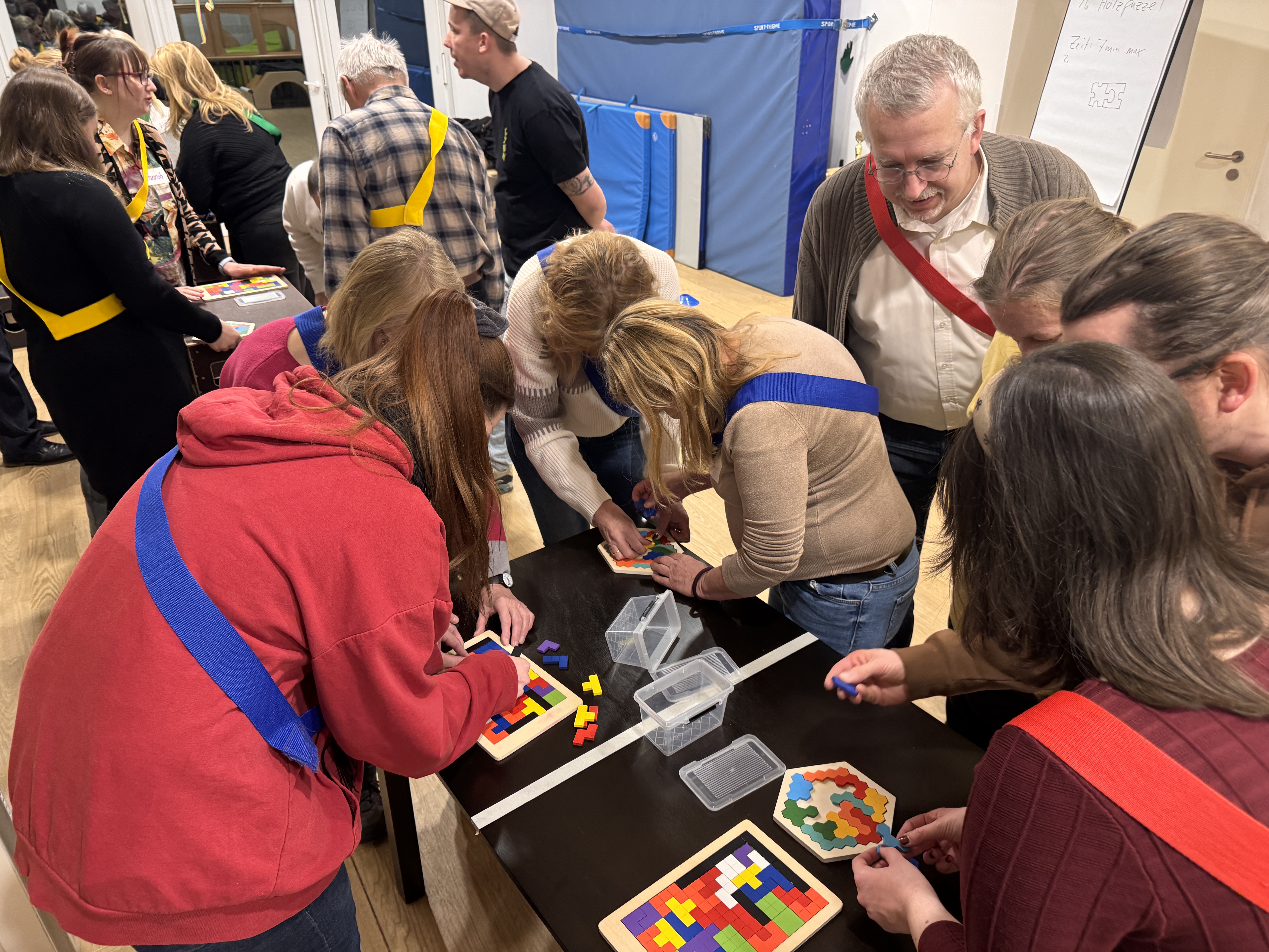 Ein Team von Erwachsenen, die farbige Schärpen tragen, arbeitet an einem Tisch in einem hell erleuchteten Raum gemeinsam an bunten Puzzlebrettern und feiert ihre Leistungen, während im Hintergrund weitere Menschen und Aktivitäten zu sehen sind.