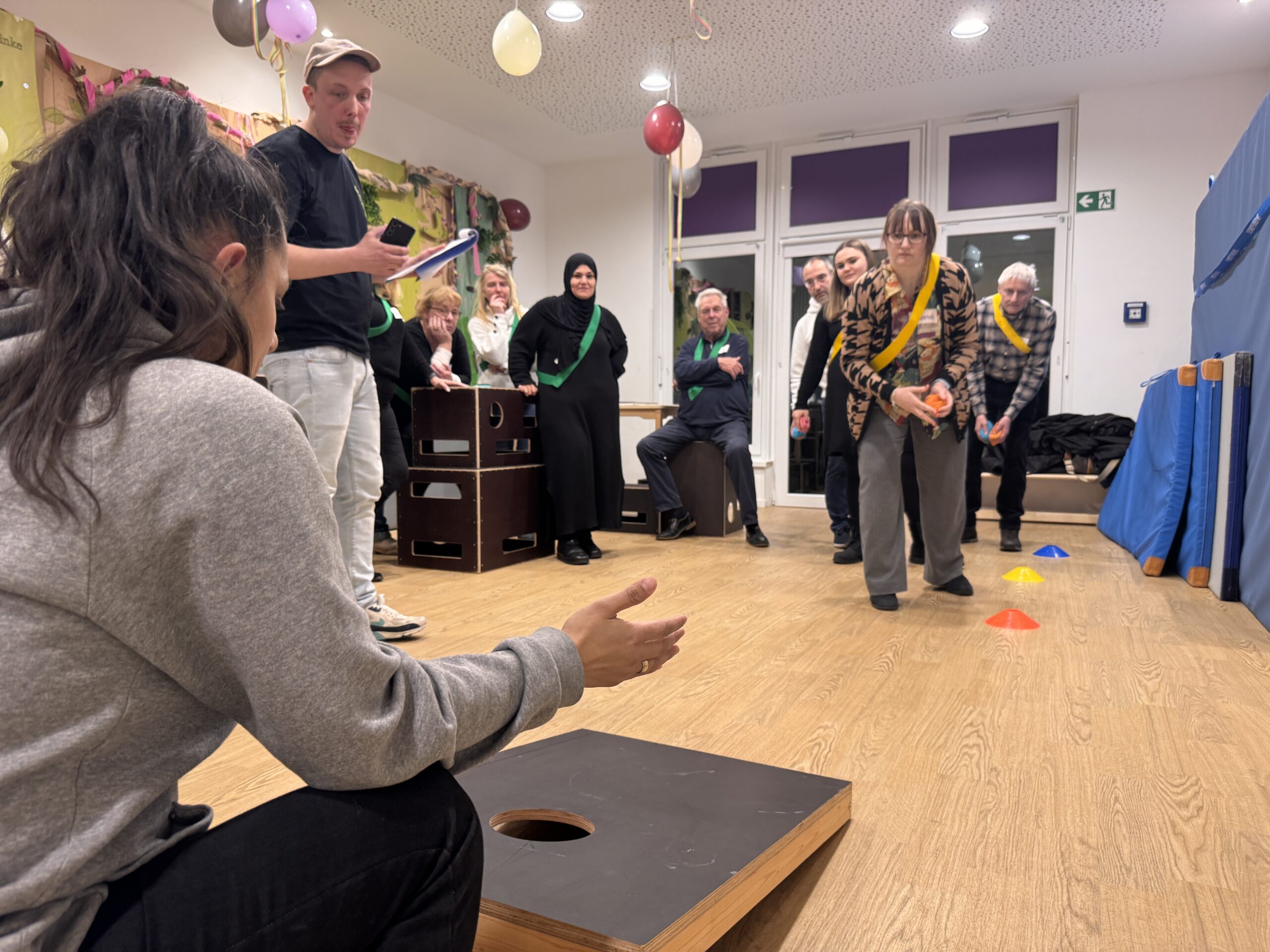 Eine Gruppe von Menschen in einem geschmückten Raum feiert, während eine Frau aus dem Team versucht, einen Sitzsack in ein Cornhole-Brett zu werfen, während eine andere Person bereit sitzt, um ihn zurückzuholen. Andere stehen in der Nähe, einige tragen Schärpen, lächeln und genießen die Feierstimmung.
