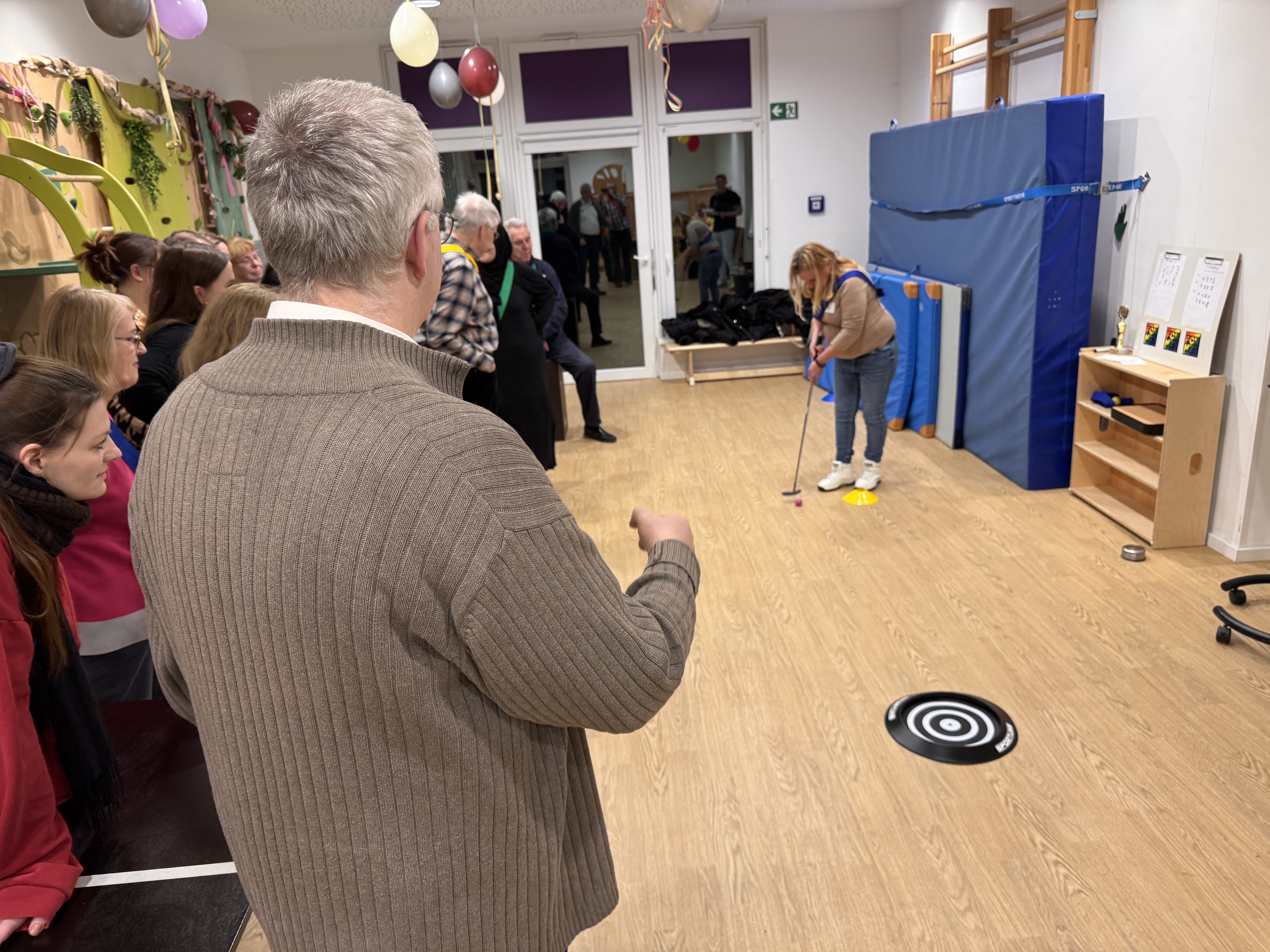 Eine Gruppe Erwachsener feiert, während eine Frau, die von ihrem Team angefeuert wird, mit einem kleinen Stock ein Indoor-Zielspiel auf einem Holzboden spielt. Der Raum ist mit Luftballons geschmückt, und auf dem Boden liegt eine Zielmatte - perfekt zum gemeinsamen Feiern.