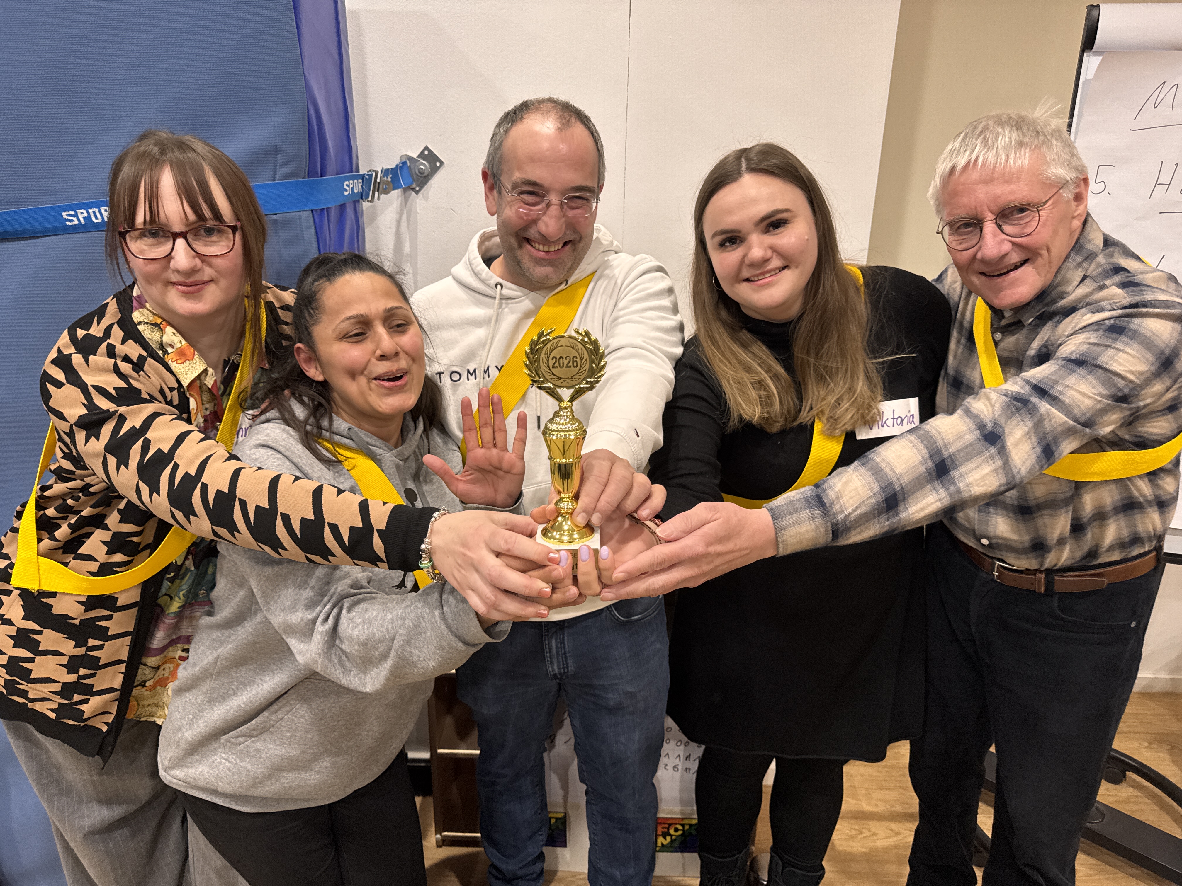 Fünf lächelnde Erwachsene stehen dicht beieinander, jeder hält einen kleinen goldenen Pokal mit der Aufschrift 2023. Die Mannschaft trägt gelbe Schärpen und Namensschilder und versammelt sich, um ihre Leistung zu feiern und einen gemeinsamen Sieg zu bejubeln.