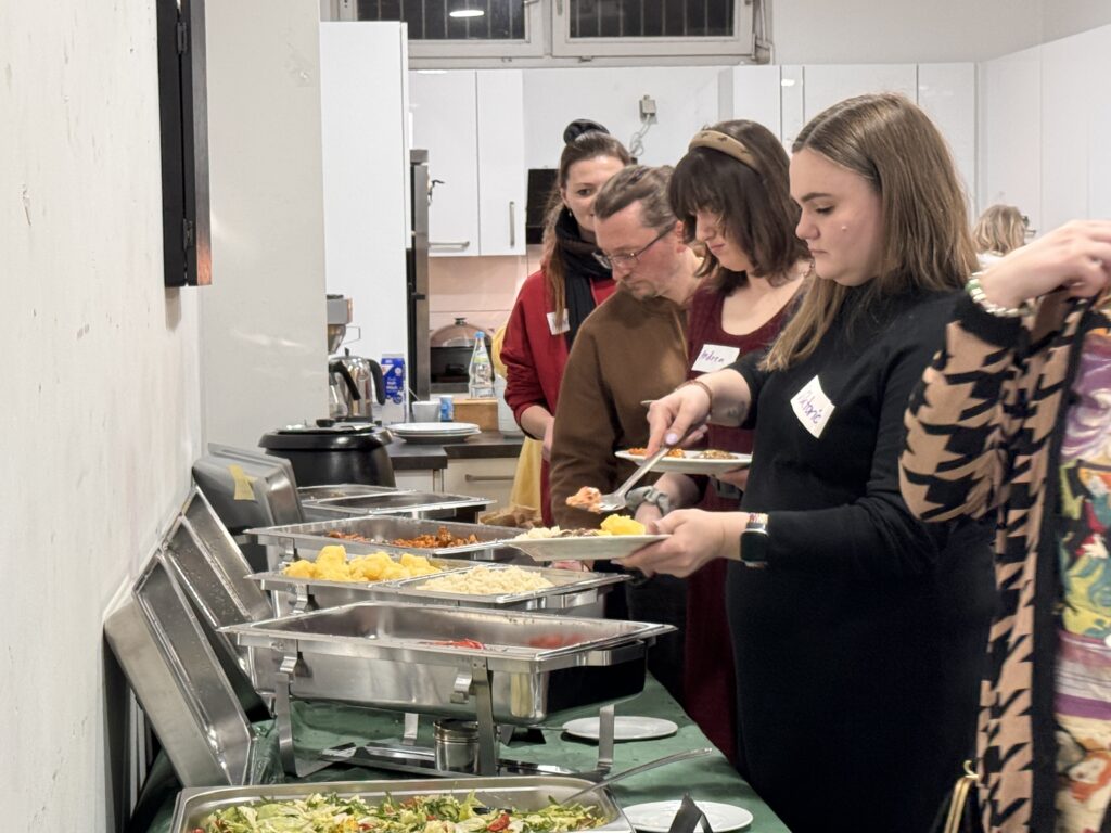 Eine Gruppe von Personen, wahrscheinlich ein Team, steht in einer Reihe an einem Buffet und bedient sich aus Tabletts. Sie scheinen bei einer Veranstaltung in einem Innenraum zu feiern, jeder hält einen Teller und trägt ein Namensschild in einem hellen Küchenbereich.