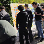 Bei einem lebhaften Graffiti-Workshop steht eine Gruppe von Menschen im Freien vor großen weißen Tafeln und sprüht bunte Graffiti. Die sonnige Szene im Grünen fängt die Konzentration und den Erfolg der Teilnehmer auf ihr Kunstwerk ein.