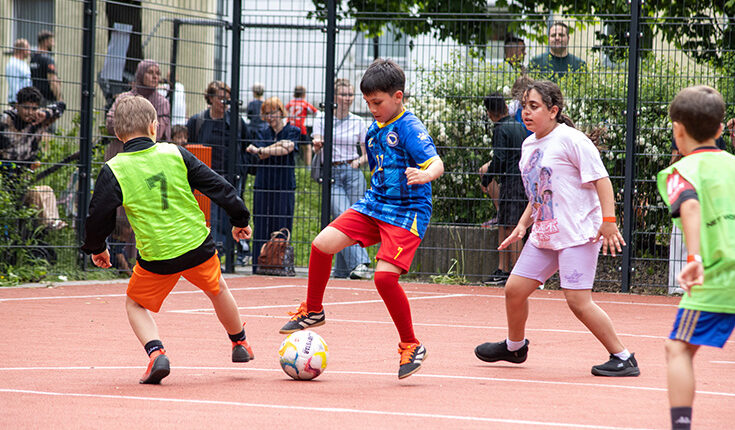Vier Kinder spielen Fußball auf einem Platz im Freien, während die Leute hinter einem Zaun zusehen. Ein Junge in einem blauen Trikot kontrolliert den Ball, bereit, ihn an den Verteidigern in grünen, schwarzen und weißen Trikots vorbei zu schießen, und träumt vom Ruhm der Thermo 2026.